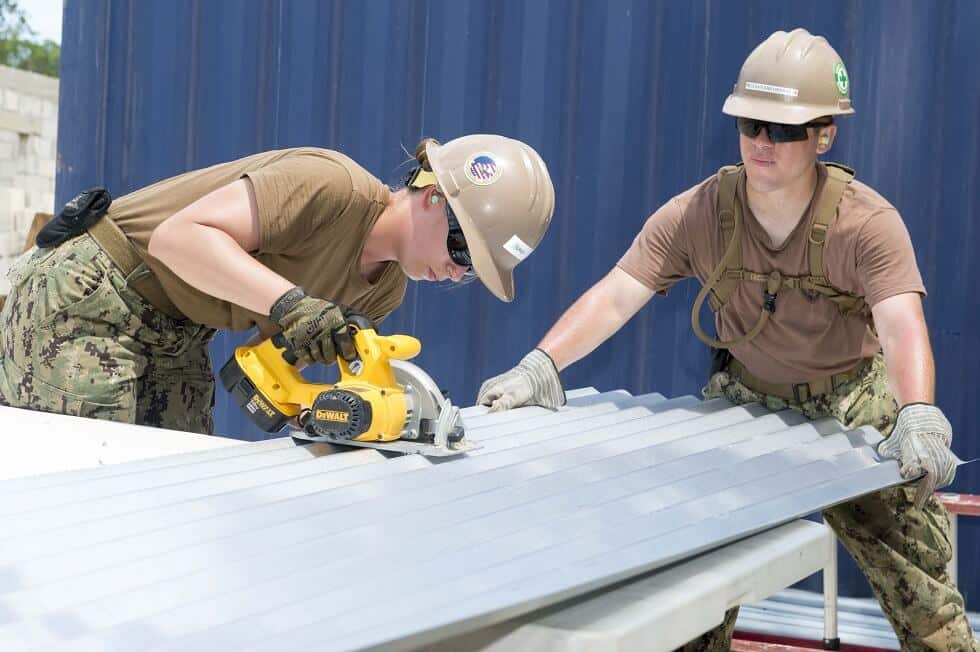 Two workers in hard hats cutting a corrugated metal roofing panel with a circular saw