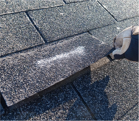 Gloved hand pointing to a damaged, granule-worn asphalt shingle during a roof inspection