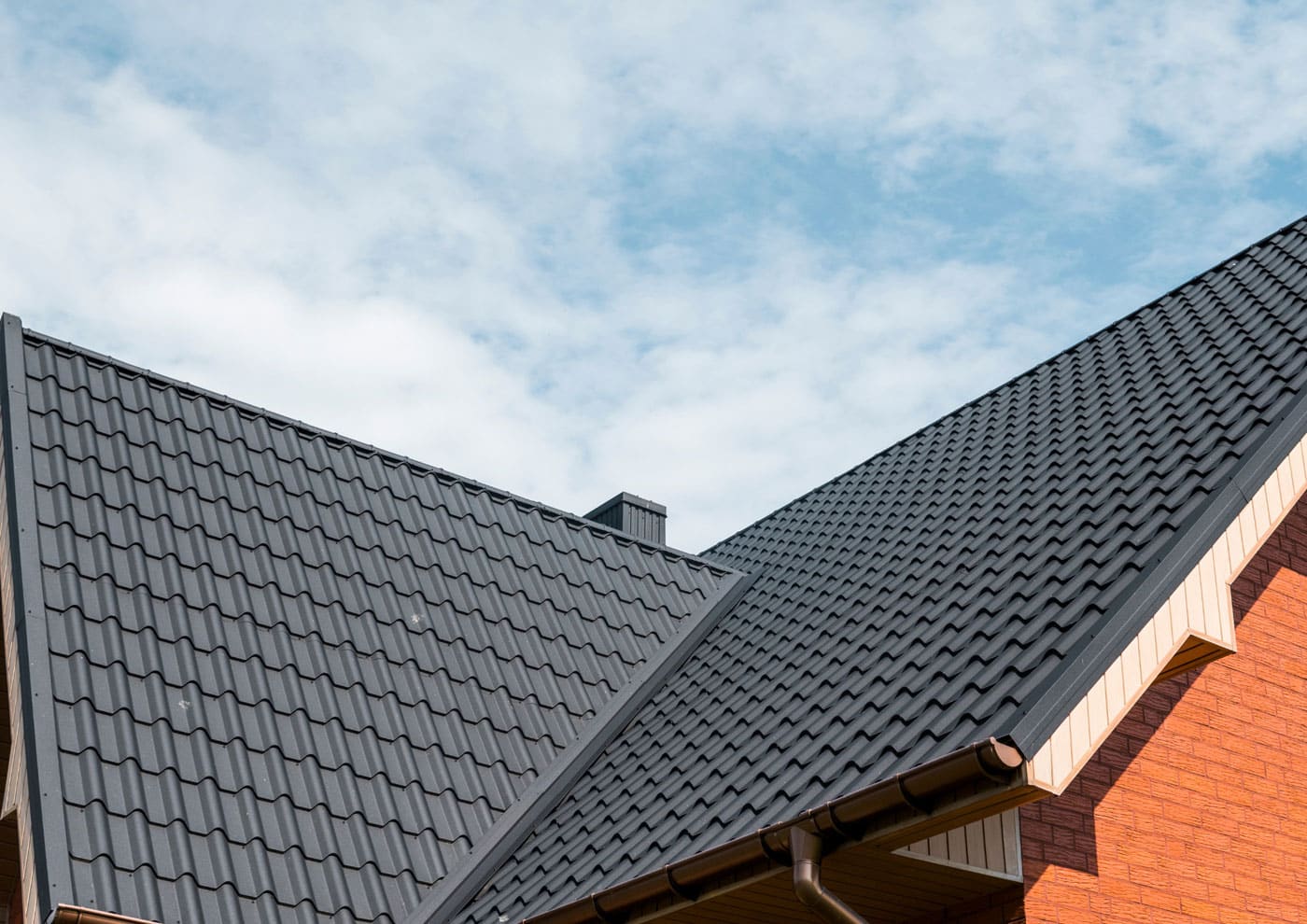Dark gray tile-profile roof panels on a brick house against a cloudy sky