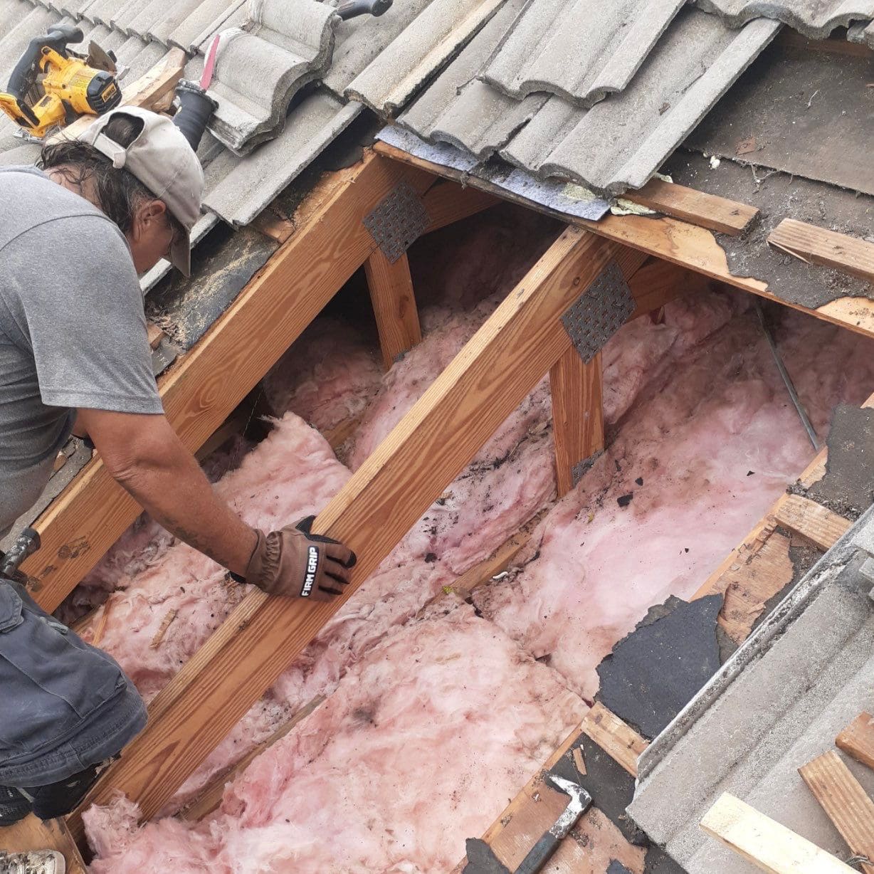 Roofer repairing wood rafters under a removed section of concrete tile roof, insulation visible below