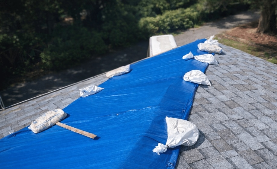 Blue emergency tarp secured with sandbags over a damaged gray shingle roof